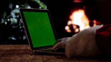 Kırmızı takım elbiseli ve beyaz eldivenli Noel Baba, şöminenin önünde yeşil ekran krom anahtarı ve Noel ağacı olan bir dizüstü bilgisayarda çalışır. Dijital ya da temalı planlar için sıcak bir festival atmosferi.