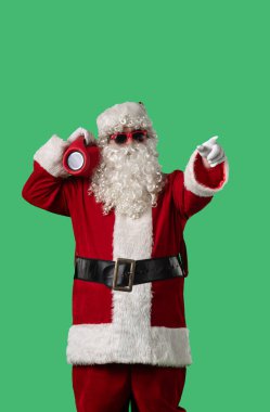 Beyaz kıvırcık sakallı, kırmızı takım elbiseli ve güneş gözlüklü Noel Baba yeşil arka planda taşınabilir bir hoparlörle neşeyle dans ediyor. Noel temalı kompozisyonlar için mükemmel.