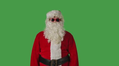 Beyaz kıvırcık sakallı, kırmızı takım elbiseli ve güneş gözlüklü Noel Baba yeşil arka planda gizli bir işaret yapıyor. Noel temalı kompozisyonlar ve tatil düzeni için ideal.