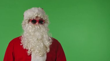 Beyaz kıvırcık sakallı, kırmızı takım elbiseli ve güneş gözlüklü Noel Baba yeşil arka planda gizli bir işaret yapıyor. Noel temalı kompozisyonlar ve tatil düzeni için ideal.