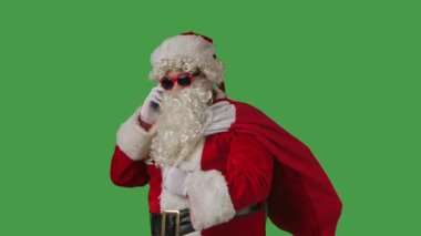 Beyaz kıvırcık sakallı Noel Baba, kırmızı takım elbise ve güneş gözlüğü takıyor, hediye dolu bir çantayla yeşil arka planda akıllı telefondan konuşuyor. Noel temalı kompozisyonlar için mükemmel.