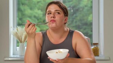 Masada oturan bir kadın beyaz bir kaseden yemek yiyor. Arka planda yeşil bir pencere var..