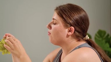 Bir kadın, sağlıklı beslenme ve yaşam tarzını vurgulayarak minimalist bir ortamda bir demet yeşil üzümden hoşlanır..