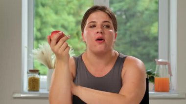 Gri kolsuz tişörtlü bir kadın, parlak ve havadar bir odada masada otururken elma yer..