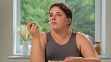 Bir kadın mutfakta oturmuş meyve salatası yiyor. Tombul bir kadın yaz sabahı sağlıklı bir vejetaryen kahvaltısının tadını çıkarır..