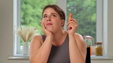 Büyük bir pencerenin kenarında oturan bir kadın, arka planda yeşillik ve rahat bir şekilde havuç tutuyor..