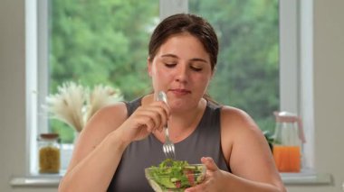 Gri kolsuz tişörtlü bir kadın evde pencerenin önünde oturmuş sağlıklı bir yemeğin tadını çıkarırken taze salata yiyor..