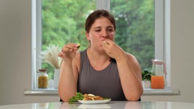 Bir kadın mutfak masasında oturmuş otlarla haşlanmış yumurtaların tadını çıkarıyor, rahat bir evde sağlıklı bir yaşam tarzı sergiliyor..