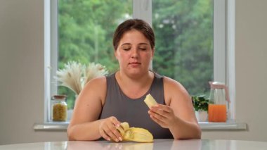 Bir kadın mutfak masasında oturmuş kahvaltısını yapıyor, taze ve lezzetli bir muz yiyor. Sağlıklı, çevre dostu bir vejetaryen kahvaltısı..