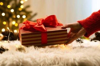 Bir el, yumuşak bir kürk halıya sarılı kırmızı bir Noel hediyesi koyar. Yılbaşı ağacındaki parıldayan tatil ışıklarının arka planına. Sıcaklık ve kutlama hissi uyandırır..