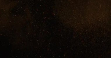 Altın tozunun soyut parçacıkları havada süzülür, koyu sarı bir zemin üzerinde izole edilmiş, rüya gibi ve sinematik bir atmosfer yaratır. Yavaş çekim.