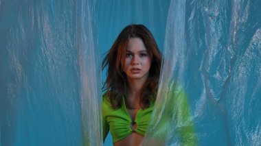 Yeşil bluzlu genç bir kadın modelin portresi, şeffaf plastik perdeli mavi bir arka plana karşı poz veriyor. Renk güzelliği, moda sanatı.