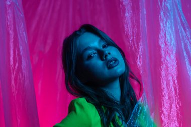 Yeşil bluzlu genç bir kadın modelin portresi, şeffaf plastik perdelerle aydınlatılmış pembe bir arka plana karşı poz veriyor. Renk güzelliği, moda sanatı.