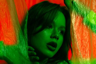 Yeşil ışıkta şeffaf plastik perdelerle aydınlatılmış kırmızı bir zemine poz veren genç bir kadın modelin portresi. Renk güzelliği, moda sanatı.