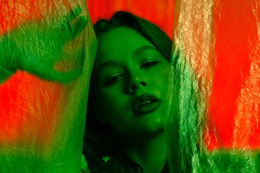 Yeşil ışıkta şeffaf plastik perdelerle aydınlatılmış kırmızı bir zemine poz veren genç bir kadın modelin portresi. Renk güzelliği, moda sanatı.