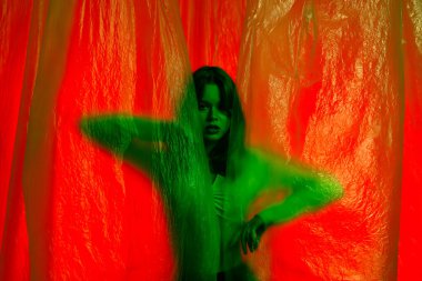Yeşil bluzlu genç bir kadın modelin portresi, şeffaf plastik perdelerle aydınlatılmış kırmızı arka planda poz veriyor. Renk güzelliği, moda sanatı.