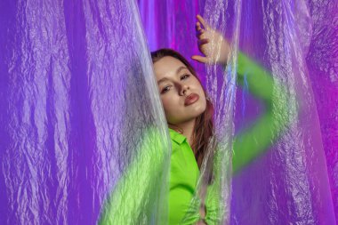 Yeşil bluzlu genç bir kadın modelin portresi, şeffaf plastik perdeli pembe ışıkla aydınlatılmış mor bir arka plana karşı poz veriyor. Renk güzelliği, moda sanatı.