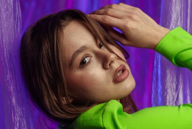 Yeşil bluzlu genç bir kadın modelin portresi, şeffaf plastik perdeli pembe ışıkla aydınlatılmış mor bir arka plana karşı poz veriyor. Renk güzelliği, moda sanatı.