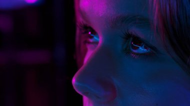 Mor ve mavi neon ışıklarla aydınlatılmış bir kadının yüzüne yakın çekim, dokular ve özellikler vurgulanıyor.