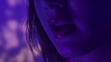 Canlı mor neon ışıkla aydınlatılan parlak dudaklar gizemli ve yansıtıcı bir ruh hali yaratıyor..