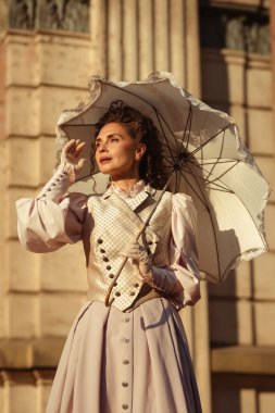 Pastel renkli Viktorya tarzı bir elbise ve eldiven giymiş zarif bir kadın dantel bir şemsiyenin altında güneşin altın ışıklarının tadını çıkarıyor. Tarihsel modanın sonsuz cazibesi ve sofistikesini somutlaştırıyor..