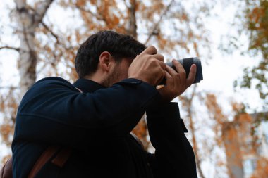 Sonbaharda elinde fotoğraf makinesi olan bir kişi, renkli yapraklarla çevrili bir parkta fotoğraf çekiyor..