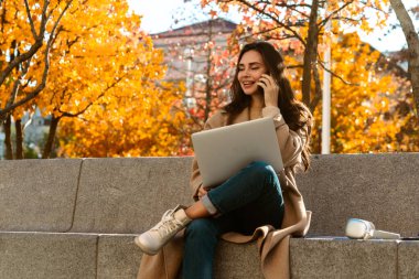Neşeli bir kadın, canlı bir sonbahar parkında bankta otururken bilgisayarında ve telefonunda birden fazla iş yapıyor..