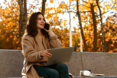 Neşeli bir kadın, canlı bir sonbahar parkında bankta otururken bilgisayarında ve telefonunda birden fazla iş yapıyor..