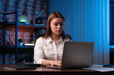 Akşamları beyaz gömlekli bir kadın dikkatle masasında dizüstü bilgisayar üzerinde çalışıyor, odak ve konsantrasyon tasvir ediyor..