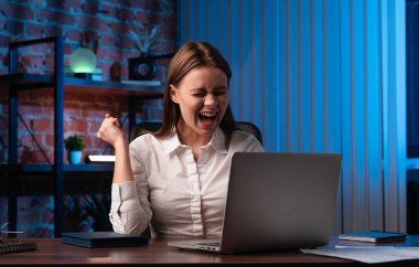 Beyaz gömlekli genç bir kadın modern ofis ortamında laptopunun başarısını kutluyor..