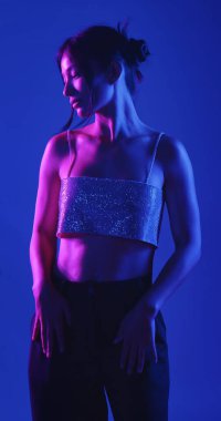 Genç bir Koreli model stüdyoda poz veriyor. Bir kadın kendine güvenerek duruyor, parlak bir bluz giyiyor, çarpıcı mavi ve pembe neon ışıklarla aydınlanıyor..