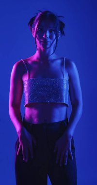 Genç bir Koreli model stüdyoda poz veriyor. Bir kadın kendine güvenerek duruyor, parlak bir bluz giyiyor, çarpıcı mavi ve pembe neon ışıklarla aydınlanıyor..