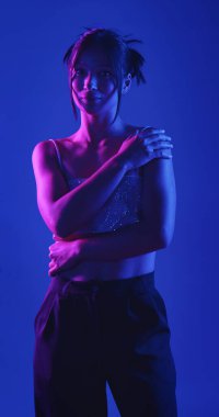 Genç bir Koreli model stüdyoda poz veriyor. Bir kadın kendine güvenerek duruyor, parlak bir bluz giyiyor, çarpıcı mavi ve pembe neon ışıklarla aydınlanıyor..