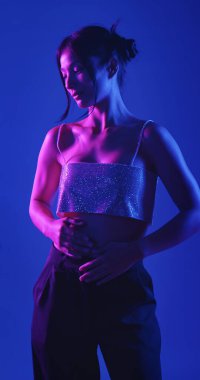 Genç bir Koreli model stüdyoda poz veriyor. Bir kadın kendine güvenerek duruyor, parlak bir bluz giyiyor, çarpıcı mavi ve pembe neon ışıklarla aydınlanıyor..