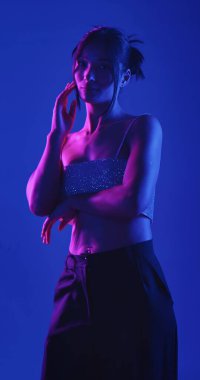 Genç bir Koreli model stüdyoda poz veriyor. Bir kadın kendine güvenerek duruyor, parlak bir bluz giyiyor, çarpıcı mavi ve pembe neon ışıklarla aydınlanıyor..