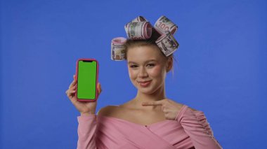 Pembe bluzlu bir kadın, saçlarında mavi arka planda banknot şeklinde bigudiler, yeşil ekranlı bir akıllı telefon tutuyor ve işaret parmağıyla işaret ediyor..