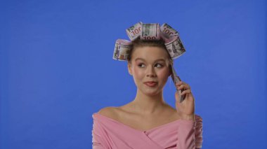 Pembe bluzlu bir kadın, saçında banknot şeklinde bukleler, mavi arka planda, bir yığın banknotla konuşur, sanki akıllı bir telefonmuş gibi.