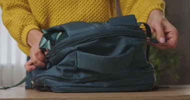 Ahşap bir masanın üzerinde koyu mavi bir sırt çantasını açan bir kadının görüntüsü. O, sarı bir örgü süveter giyiyor ve içeride. Yavaş çekim.