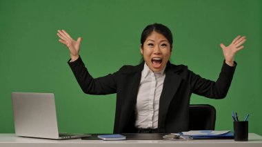 Siyah takım elbiseli genç bir Koreli iş kadını zafere ya da iyi habere sevinir. Kadın elinde laptop ve ofis malzemesi olan bir masada oturuyor ve yeşil renkli anahtar arka planda..