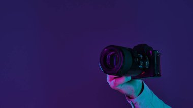 Kamerayı tutan bir elin detaylı bir yakın çekimi, parlak pembe ve mor ışıklarla aydınlatılmış, koyu bir arka plana karşı..