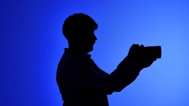 Sahne arkası profesyonel bir stüdyoda, elinde kamera tutan bir kişinin silüeti, canlı, mavi bir arkaplanda, yaratıcılığı aktarıyor..