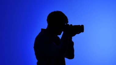 Sahne arkası profesyonel bir stüdyoda, elinde kamera tutan bir kişinin silüeti, canlı, mavi bir arkaplanda, yaratıcılığı aktarıyor..