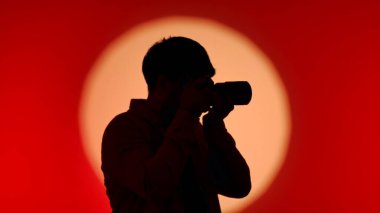 Profesyonel bir stüdyonun sahne arkasında, elinde kamera olan bir kişinin silüeti, çarpıcı kırmızı ve turuncu bir gradyan üzerine kurulmuş..