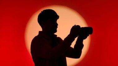 Profesyonel bir stüdyonun sahne arkasında, elinde kamera olan bir kişinin silüeti, çarpıcı kırmızı ve turuncu bir gradyan üzerine kurulmuş..