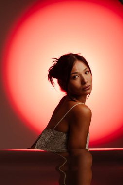 Huzurlu bir kadın zarif bir şekilde oturur, kırmızı ışıkta yıkanır, yansıması pürüzsüz bir yüzeyde görünür, dramatik ve içe dönük bir atmosfer yaratır..