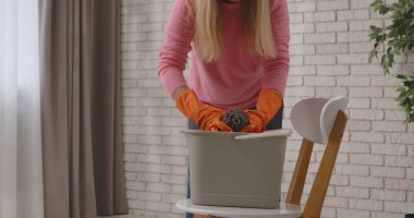 Pembe kazaklı ve turuncu eldivenli bir kadın iyi aydınlatılmış bir iç odada paspas ve kovayla temizlik yapıyor..