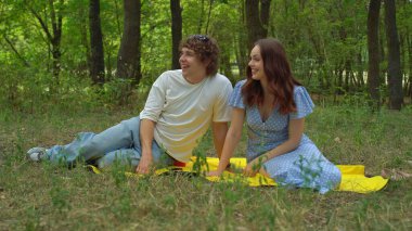Genç bir çift yemyeşil bir ormanda piknik battaniyesinde dinleniyor ve gülüyor açık havada güneşli bir öğleden sonranın tadını çıkarıyor..