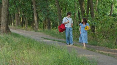 Bir adam ve bir kadın, güneşli bir günde, uzun ağaçların gölgesinde yemyeşil bir orman yolunda geziniyorlar..