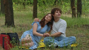 Ormanda bir battaniyenin üzerinde oturan mutlu genç bir çift ağaçlarla çevrili huzurlu bir pikniğin tadını çıkarıyor..