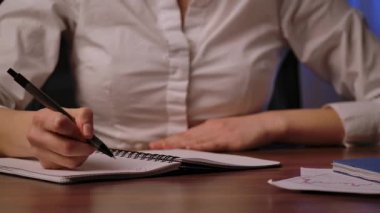 Beyaz gömlek giyen, tahta bir masanın üzerinde kalemle el yazısı yazan bir kadın..